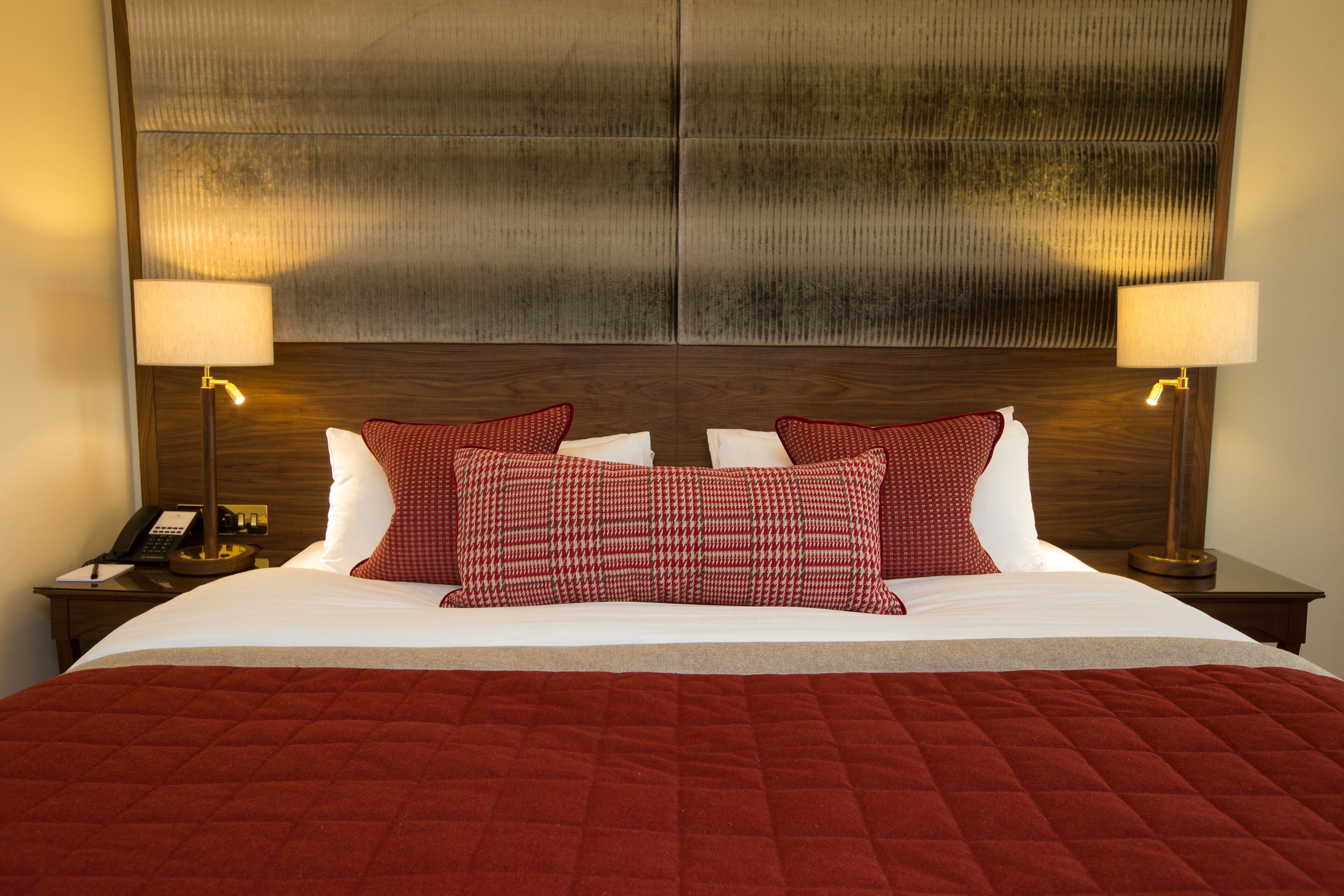 Una cama bien hecha con un edredón rojo, dos almohadas rojas estampadas y un cabecero de madera. Hay dos lámparas de noche y una mesita con un teléfono.