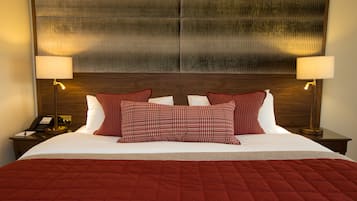 Una cama bien hecha con un edredón rojo, dos almohadas rojas estampadas y un cabecero de madera. Hay dos lámparas de noche y una mesita con un teléfono.