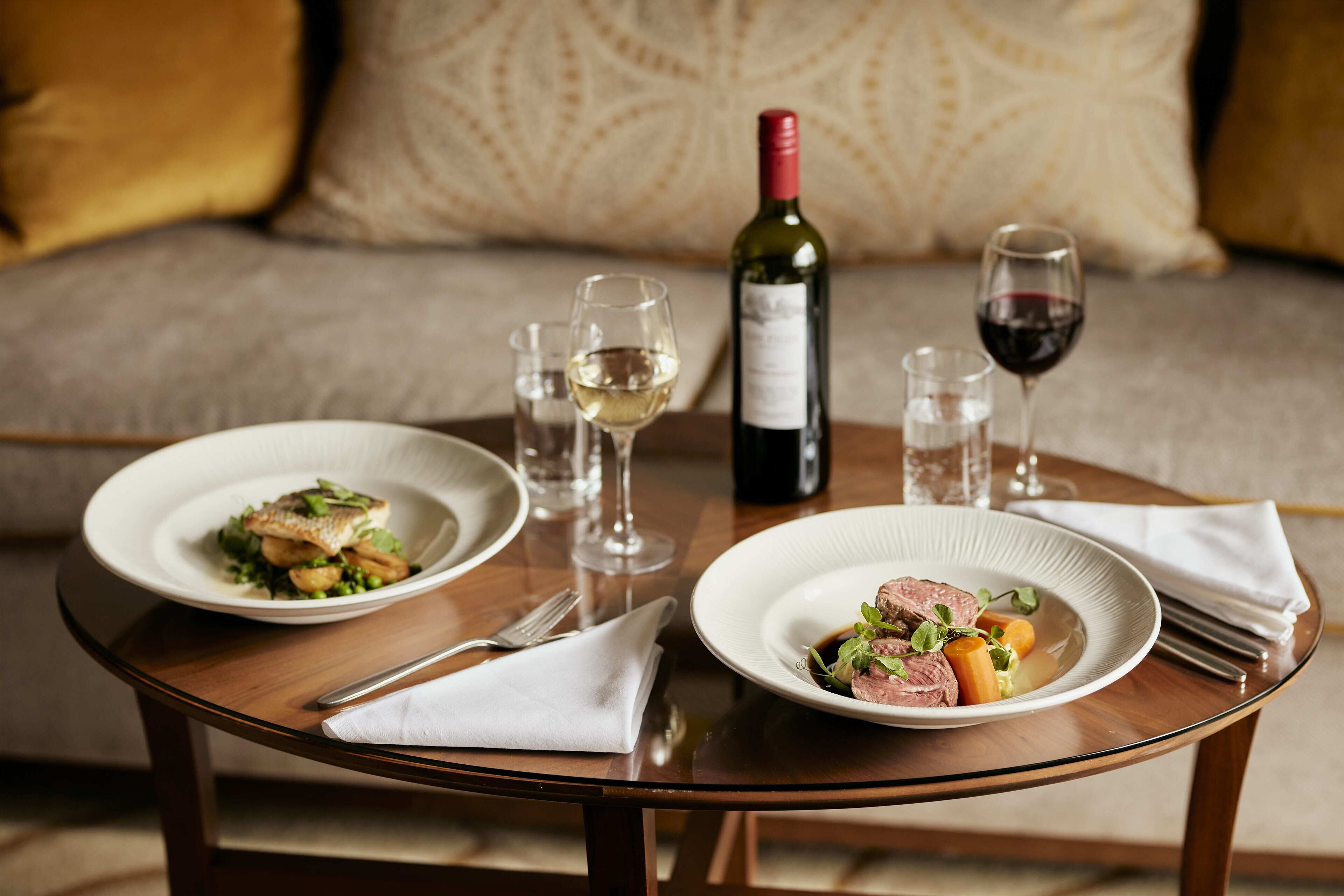 A dining table with two plates of food, a bottle of wine, and two glasses.