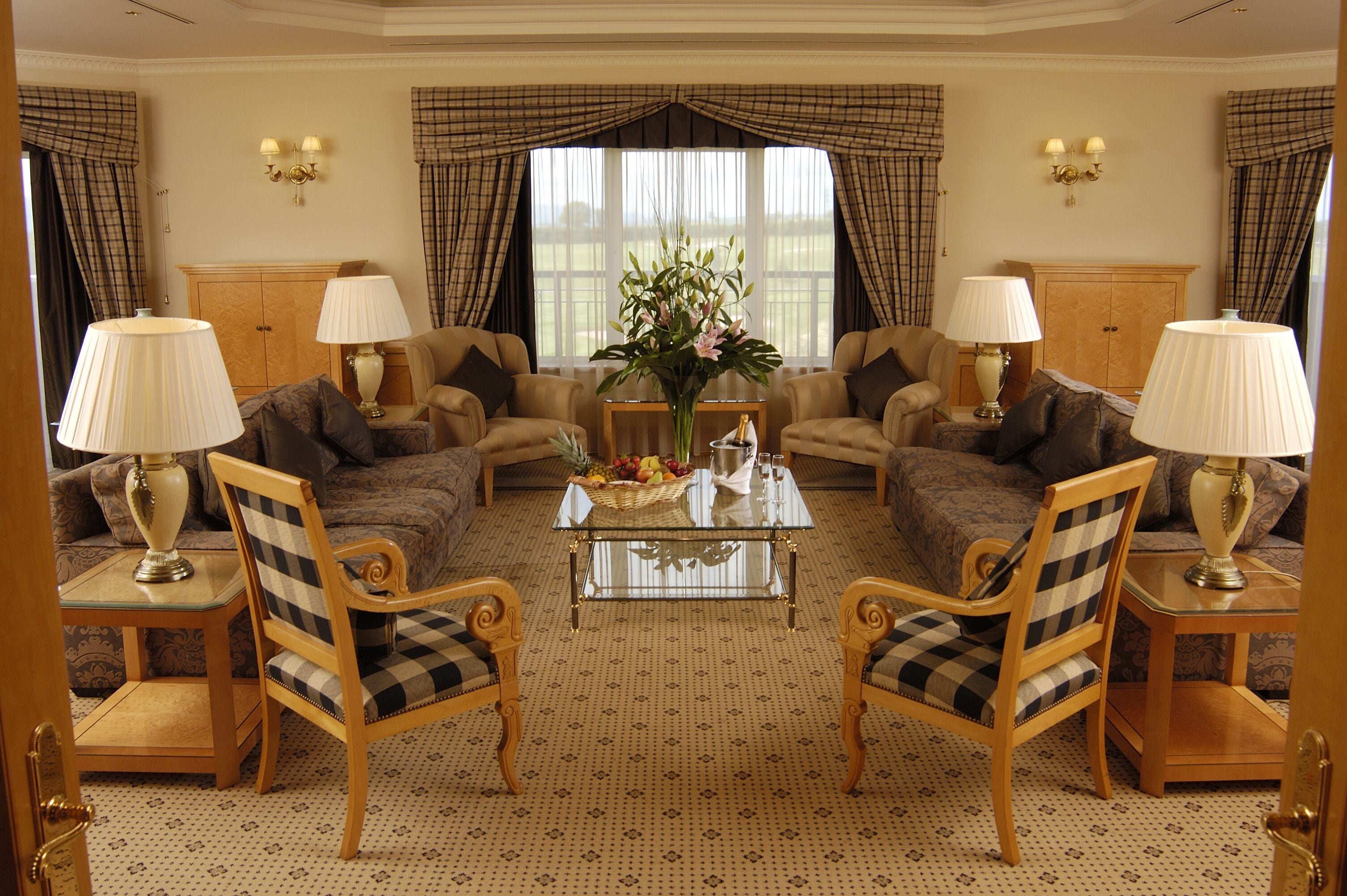 A living room with two sofas, a coffee table, and two armchairs. There are lamps on side tables, a vase with flowers on the coffee table, and a window with curtains.