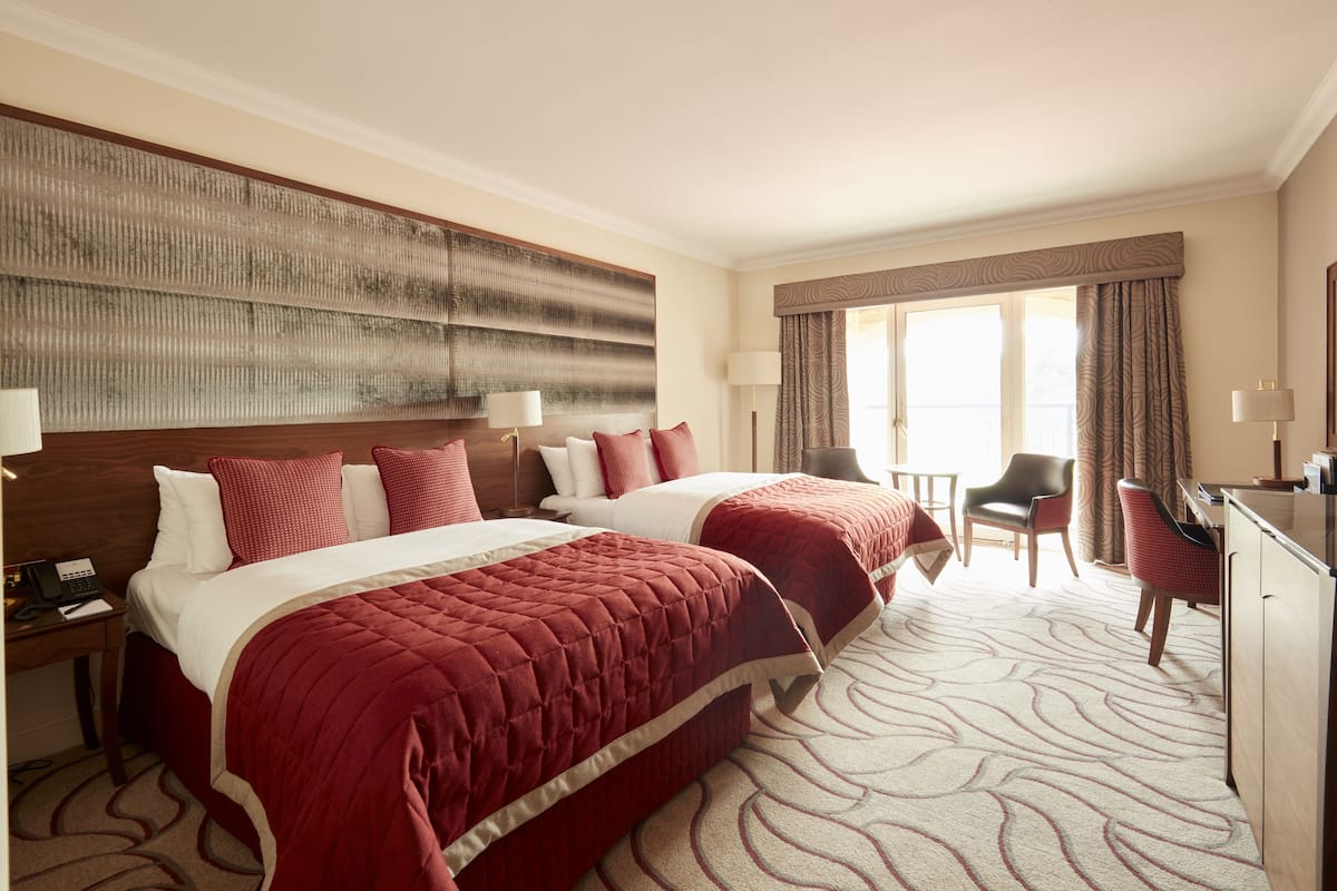 A hotel room with two beds, a desk, a chair, and a large window with curtains.