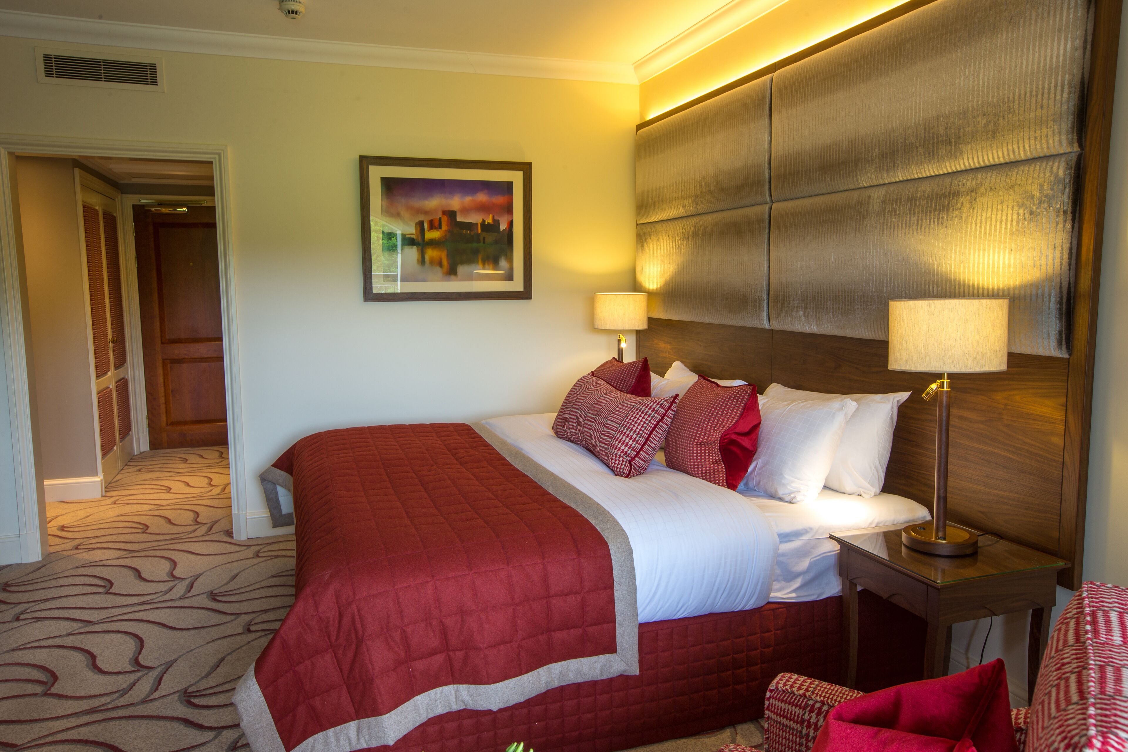 A hotel room with a large bed, two bedside tables with lamps, a headboard, a painting on the wall, and a carpeted floor.