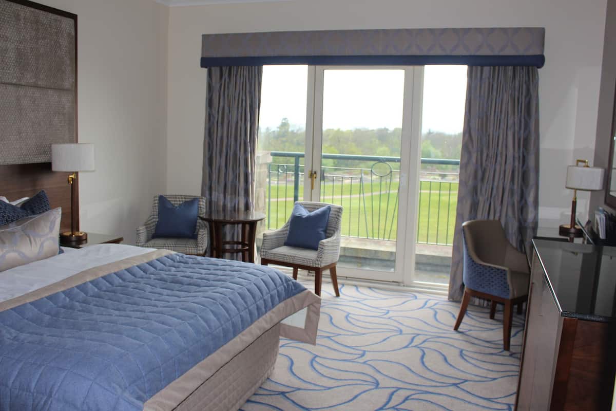 A hotel room with a large bed, two chairs, a small table, and a view of a green field through the window.
