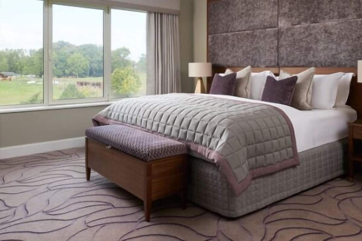 A bedroom with a large bed, two bedside tables with lamps, a bench, and a view of greenery outside.