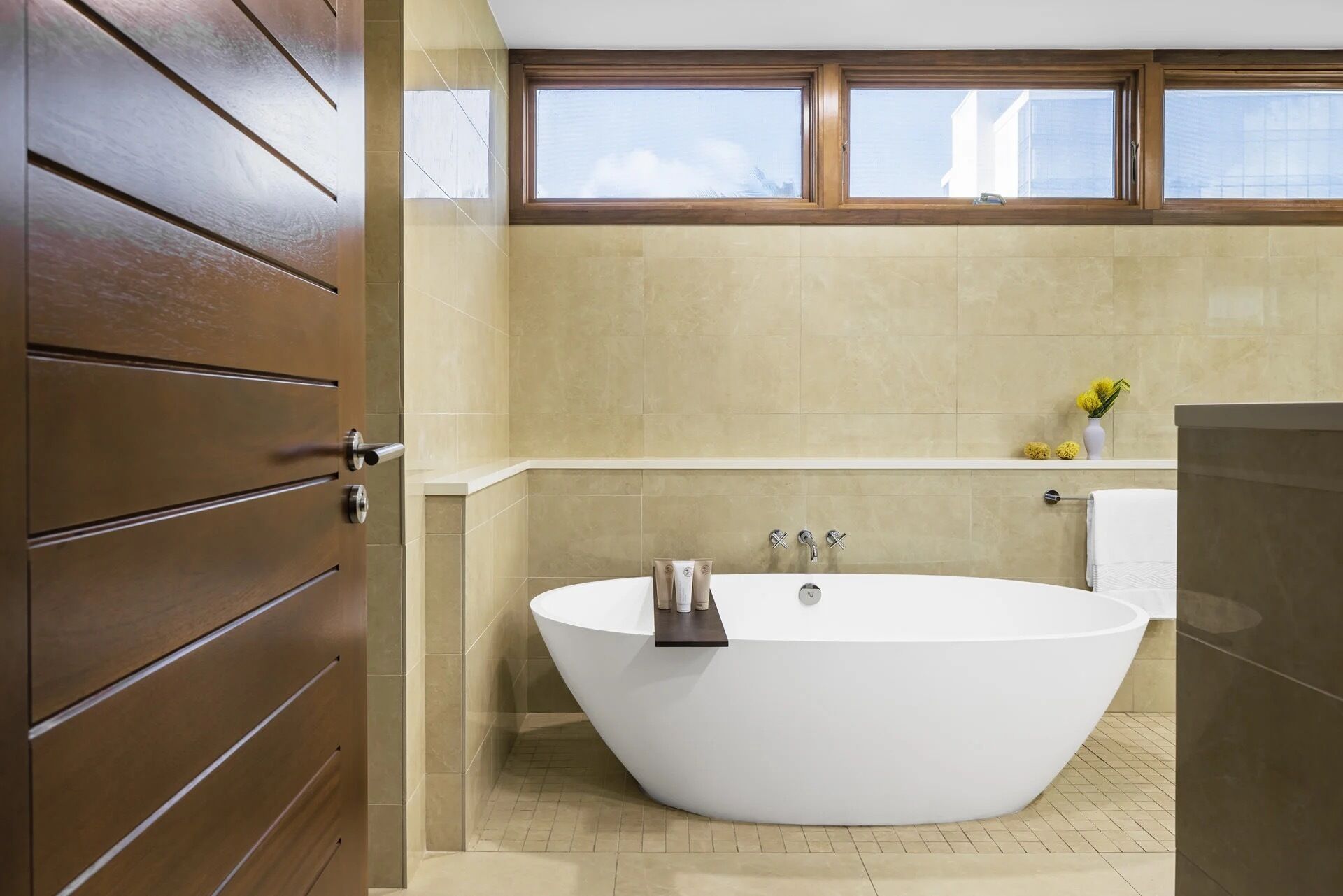 Un baño moderno con una bañera independiente, azulejos color beige, una puerta de madera y una ventana sobre la bañera.