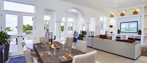 A modern dining area with a wooden table set for six, surrounded by white chairs, and a large flat-screen TV mounted on the wall.