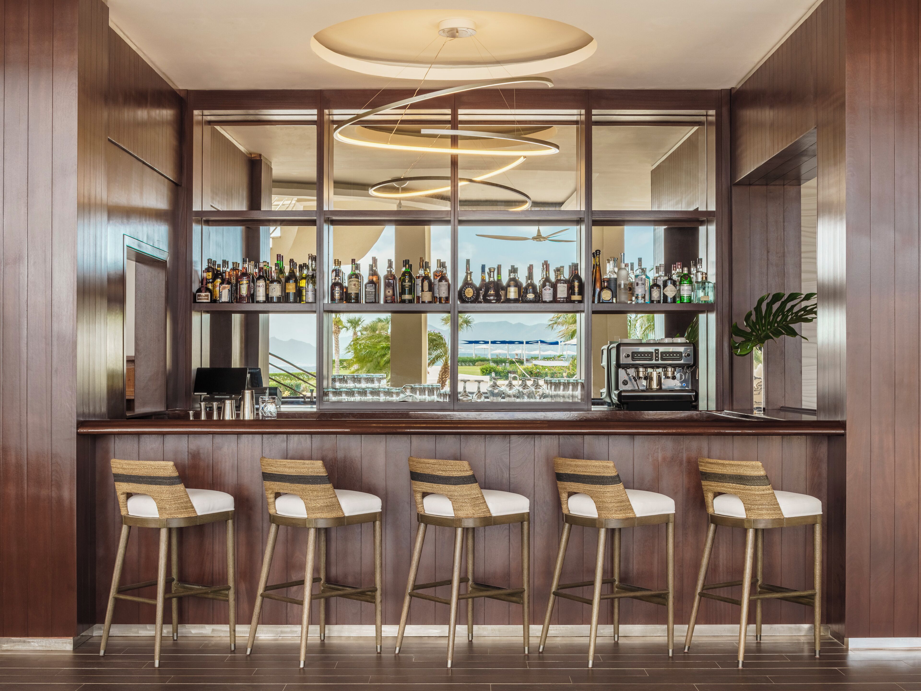 Zona de bar con paneles de madera, un ventanal con vistas a palmeras y montañas, y una lámpara moderna.