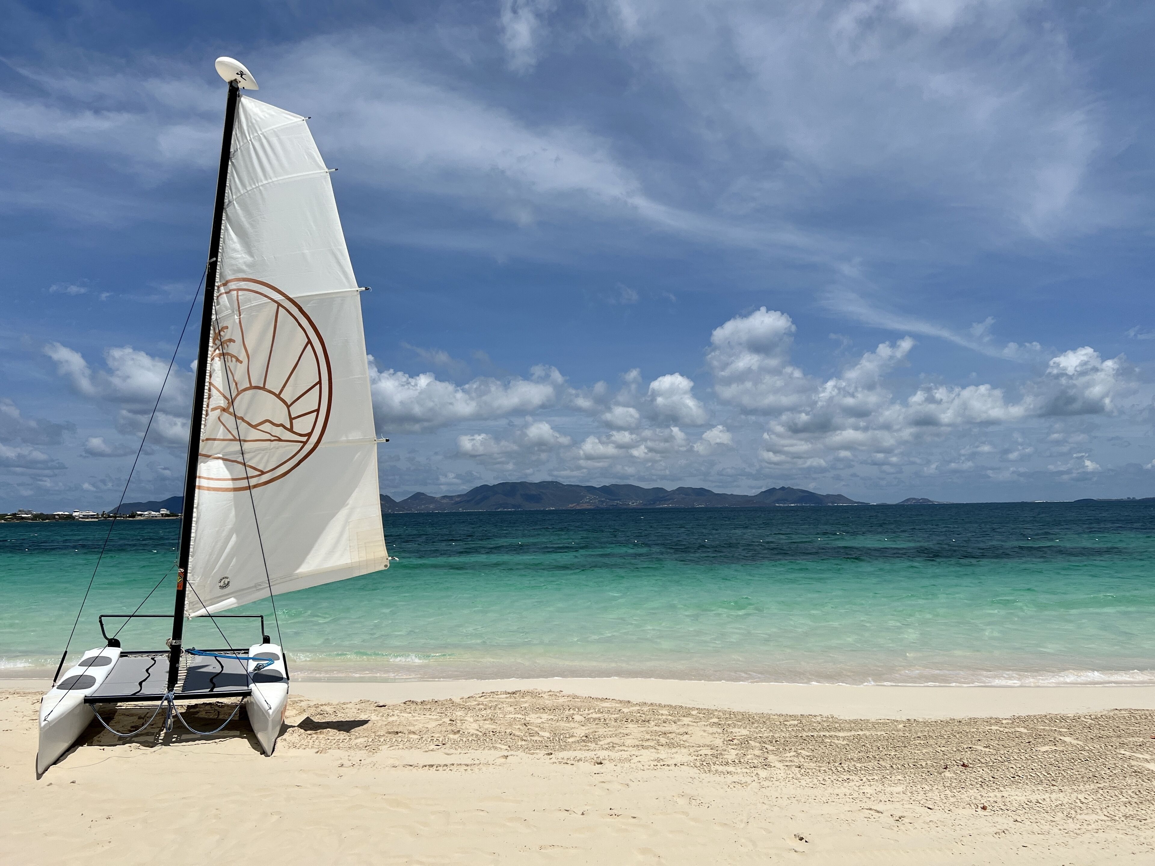 Un voilier à la voile blanche sur une plage de sable fin, avec une eau turquoise et limpide et des montagnes à l’horizon.