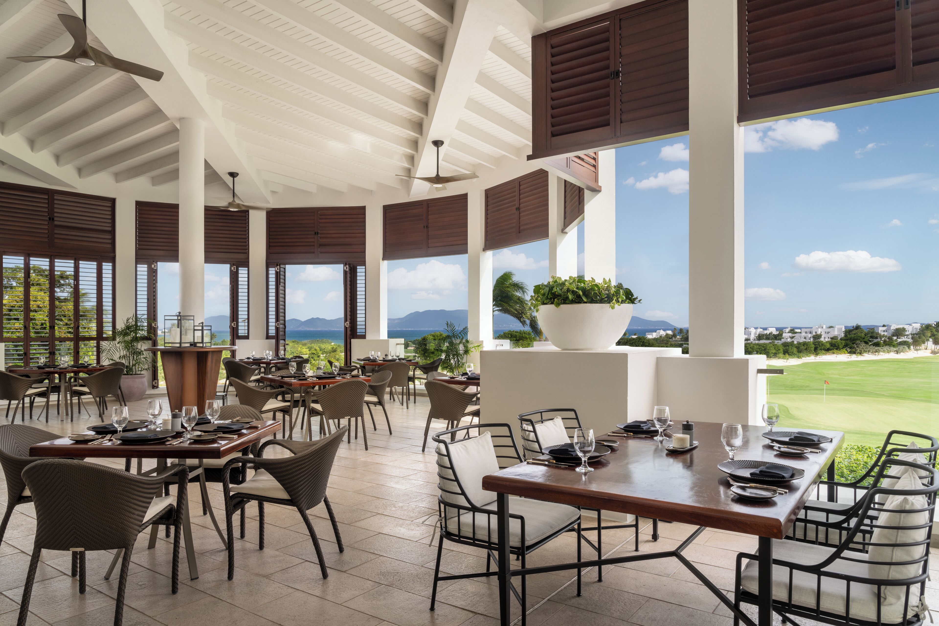 A spacious outdoor dining area with multiple tables and chairs, large windows offering a view of a golf course and mountains, and a ceiling fan.