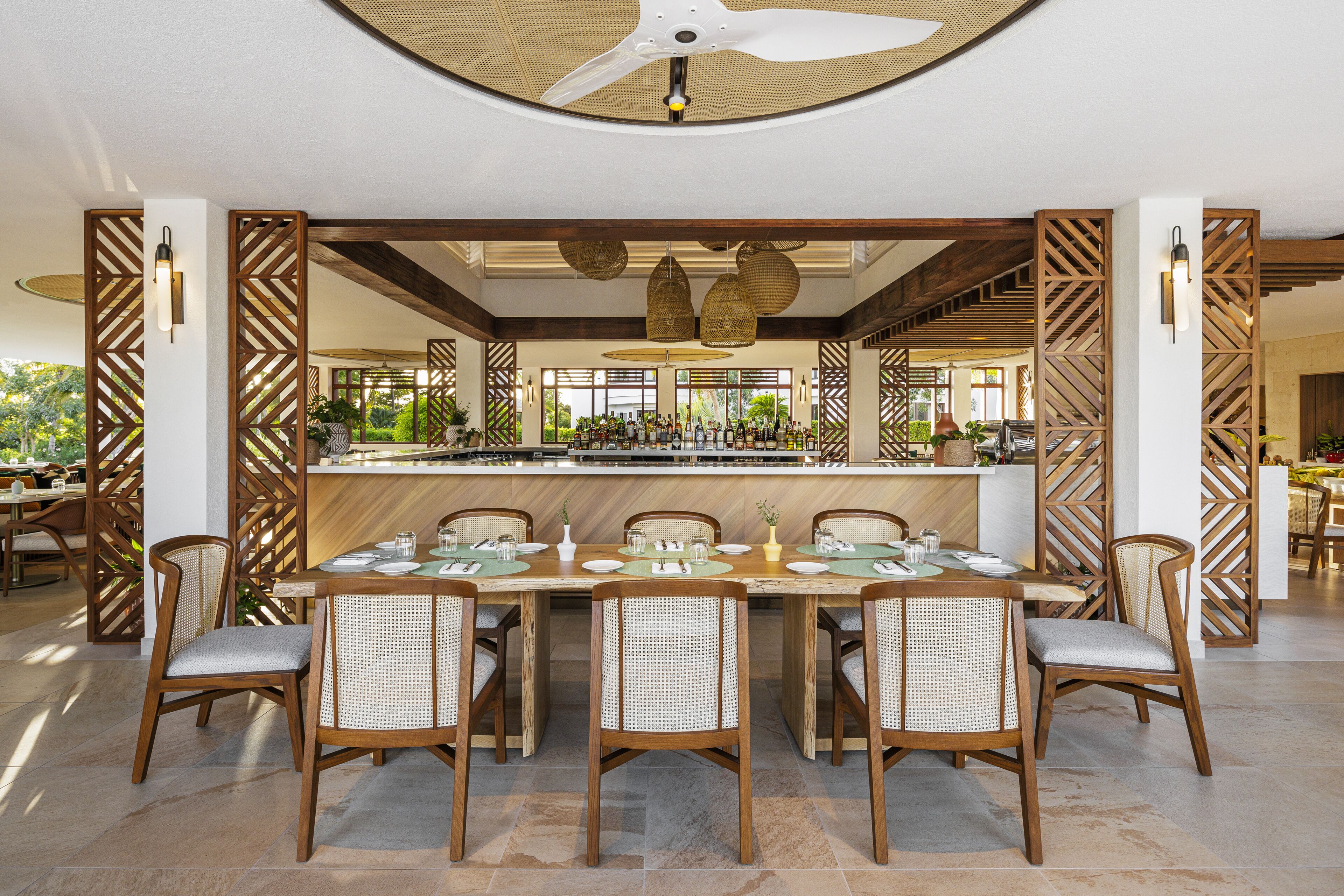 A dining area with a long table set for six, surrounded by wicker chairs, with a bar in the background.