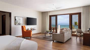 Amplia habitación de hotel con una cama grande, un sofá, una mesa de comedor y un televisor montado en la pared. La habitación tiene un balcón con vistas al océano.