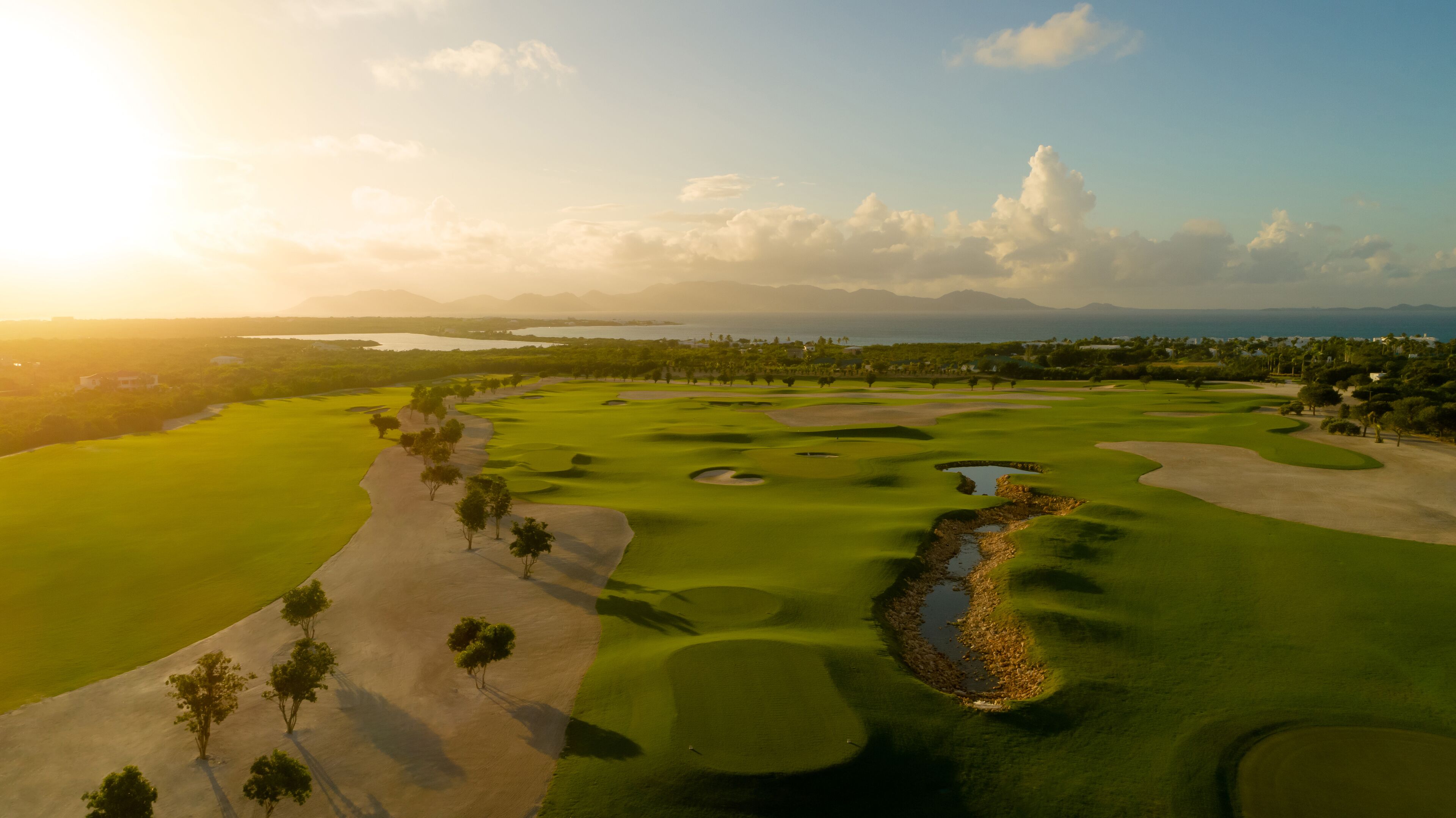 A golf course with sand bunkers, water hazards, and a clear view of the ocean and mountains in the background.