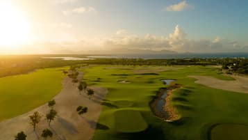 A golf course with sand bunkers, water hazards, and a clear view of the ocean and mountains in the background.