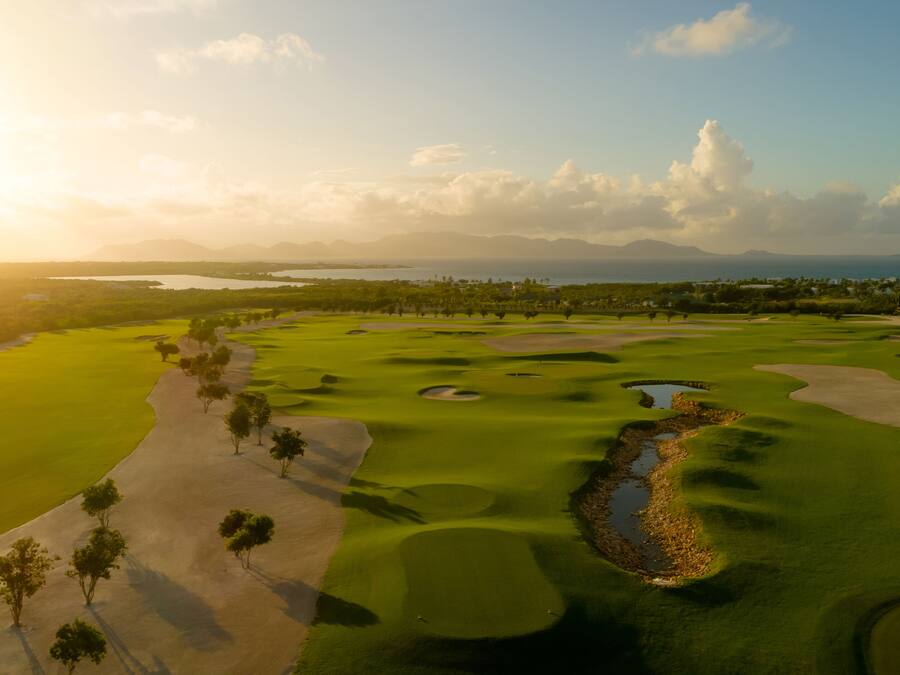 Un campo de golf con búnkeres de arena, obstáculos acuáticos y una vista despejada del océano y las montañas al fondo.