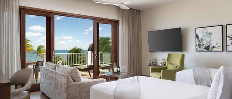 A hotel room with a bed, a sofa, a chair, a TV, and a balcony with a view of the ocean.