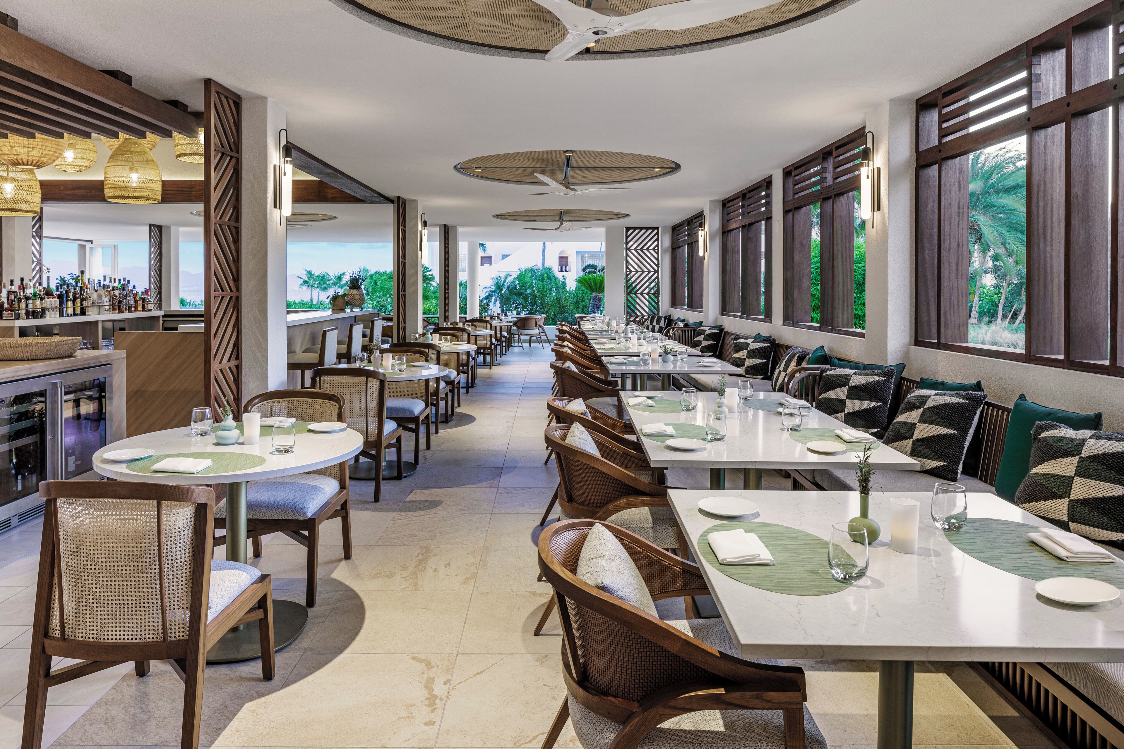 A restaurant with long communal tables, cushioned chairs, and a bar area.