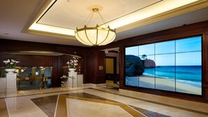 Tilava aula, jossa on suuri digitaalinäyttö, joka esittää rantamaisemaa, näyttävä kattokruunu ja marmorilattia.
