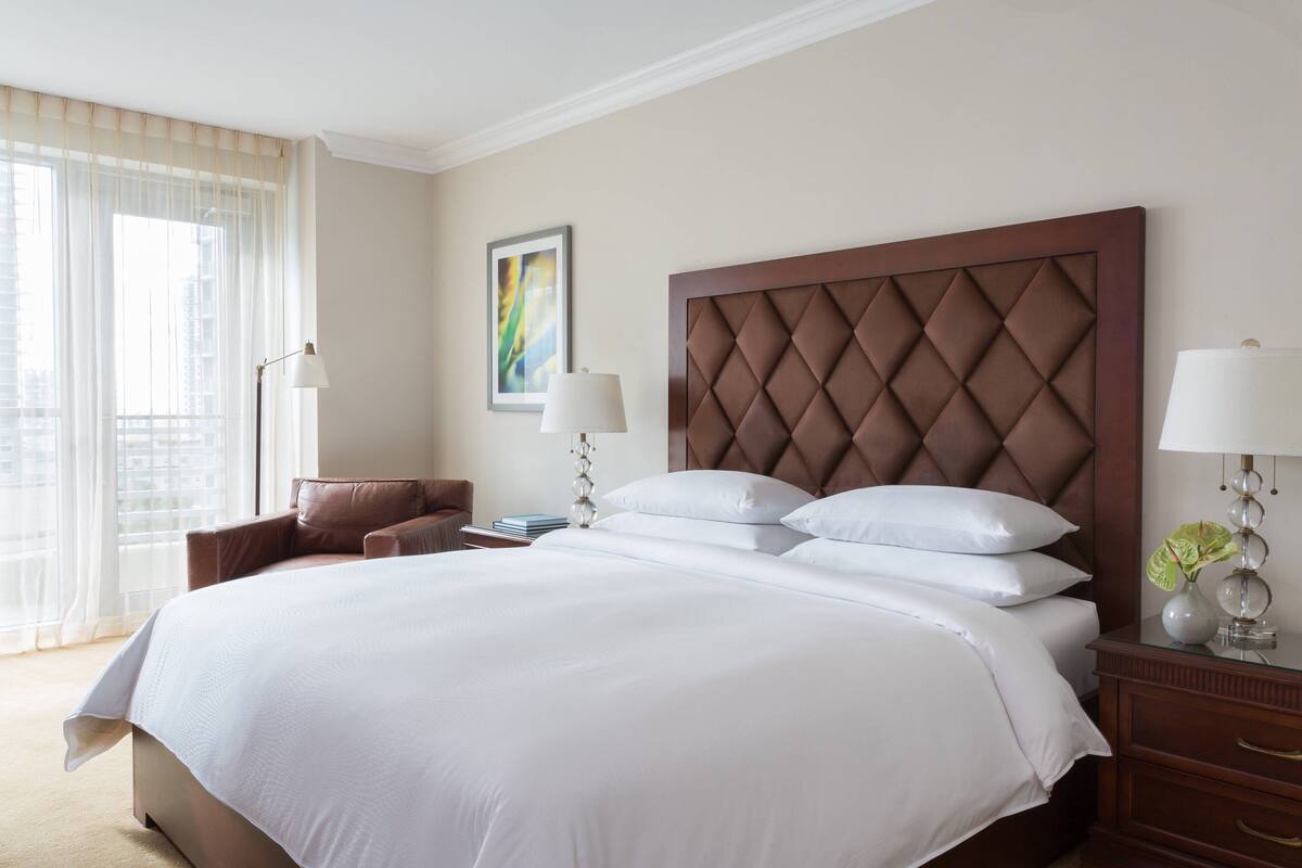 Ein Hotelzimmer mit einem großen Bett, einem Sessel, einem Nachttisch und einem Fenster mit Vorhängen.