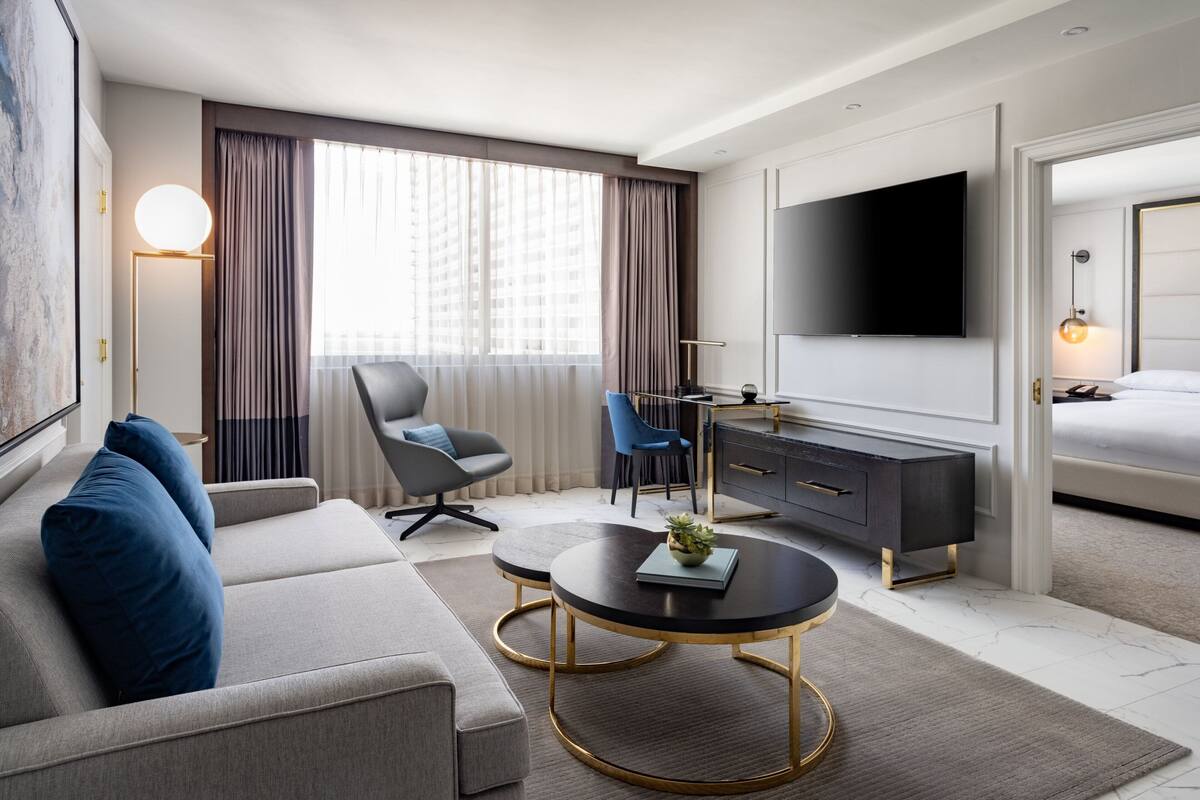 Ein modernes Hotelzimmer mit einer Couch, einem runden Couchtisch, einem Fernseher, einem Bett und einem Schreibtisch mit Stuhl.
