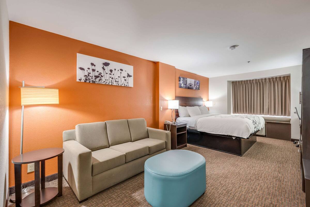 A hotel room with a sofa, a small table, a blue ottoman, a bed, and artwork on the walls.