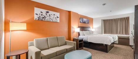 A hotel room with a sofa, a small table, a blue ottoman, a bed, and artwork on the walls.