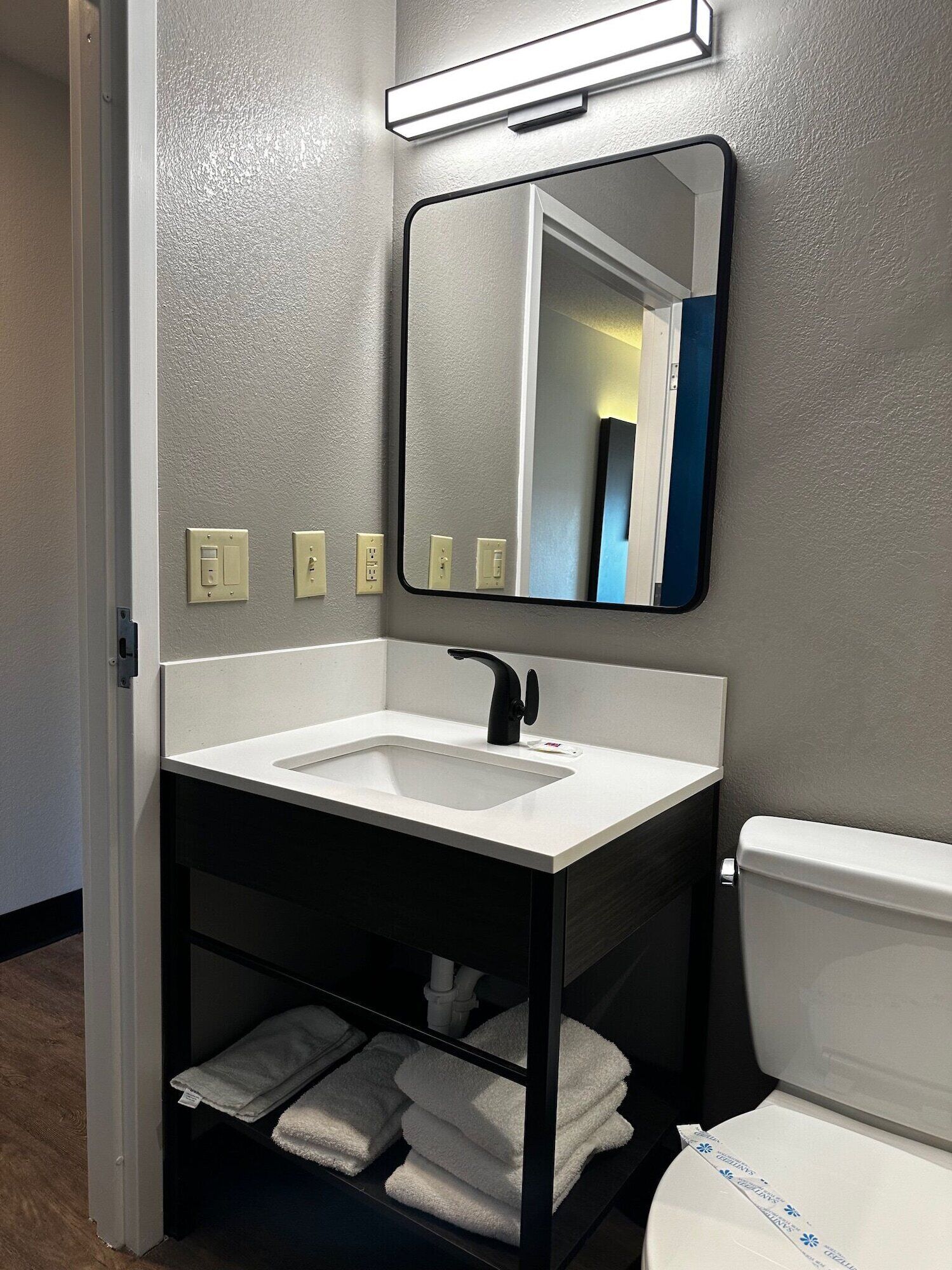 A bathroom with a white sink, black vanity, and a mirror above it. There are towels on the vanity and a toilet to the right.