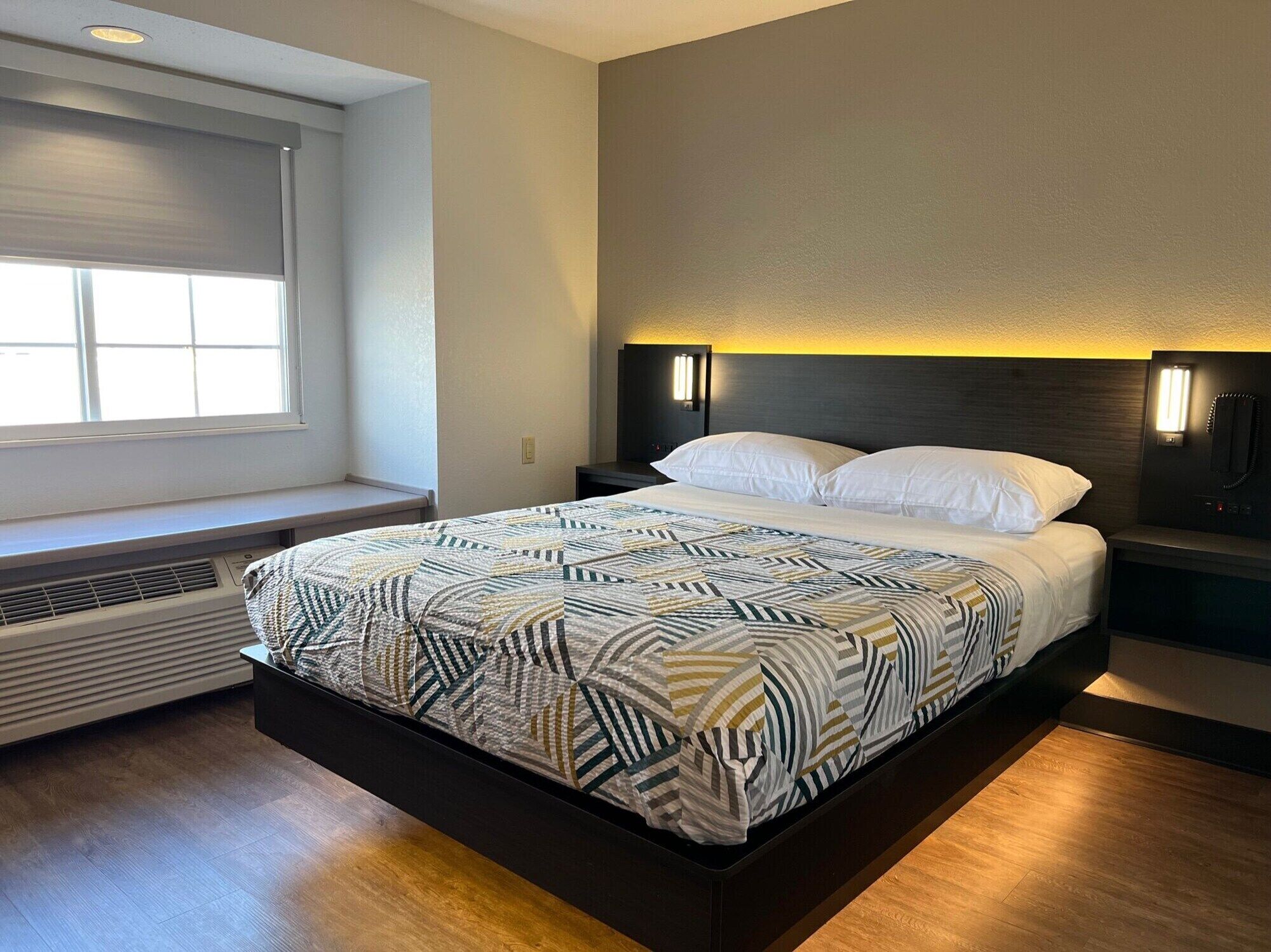 Una habitación de hotel moderna con una cama grande, una ventana con persianas, un calentador en la pared y una mesita de noche con lámpara.