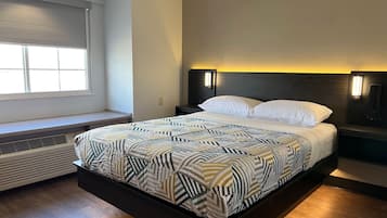 A modern hotel room with a large bed, a window with blinds, a wall-mounted heater, and a nightstand with a lamp.