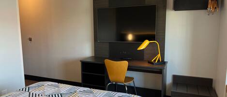 A hotel room with a bed, a desk with a TV, a chair, a bench, and a wall-mounted shelf with hooks.