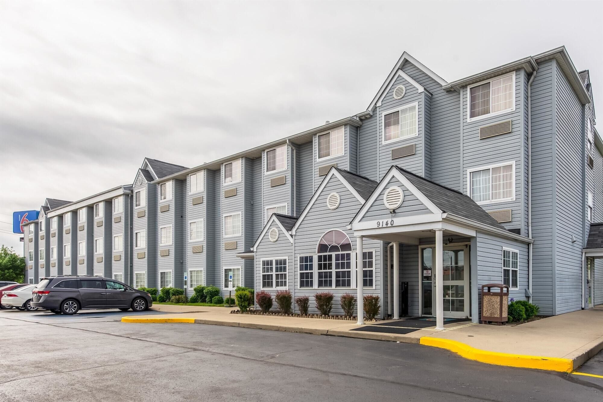 A multi-story hotel with a grey facade, a covered entrance, and a parking lot with cars.