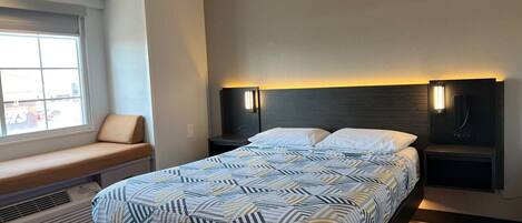 Een moderne hotelkamer met een bed, nachtkastjes, een bankje en een raam.