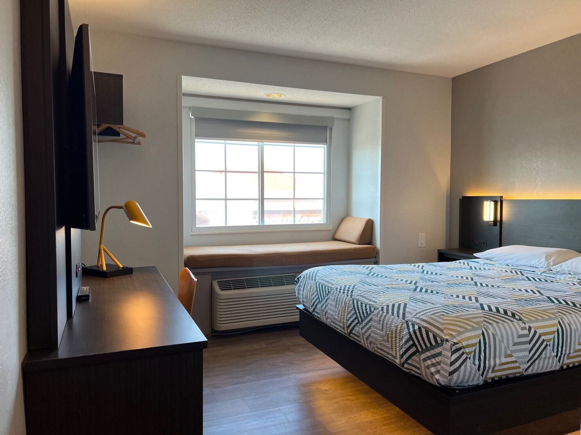 A hotel room with a bed, a desk with a lamp, a window with blinds, and a wall-mounted TV.