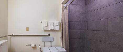 A bathroom with a toilet, a shower with a curtain, a grab bar, and a small table.