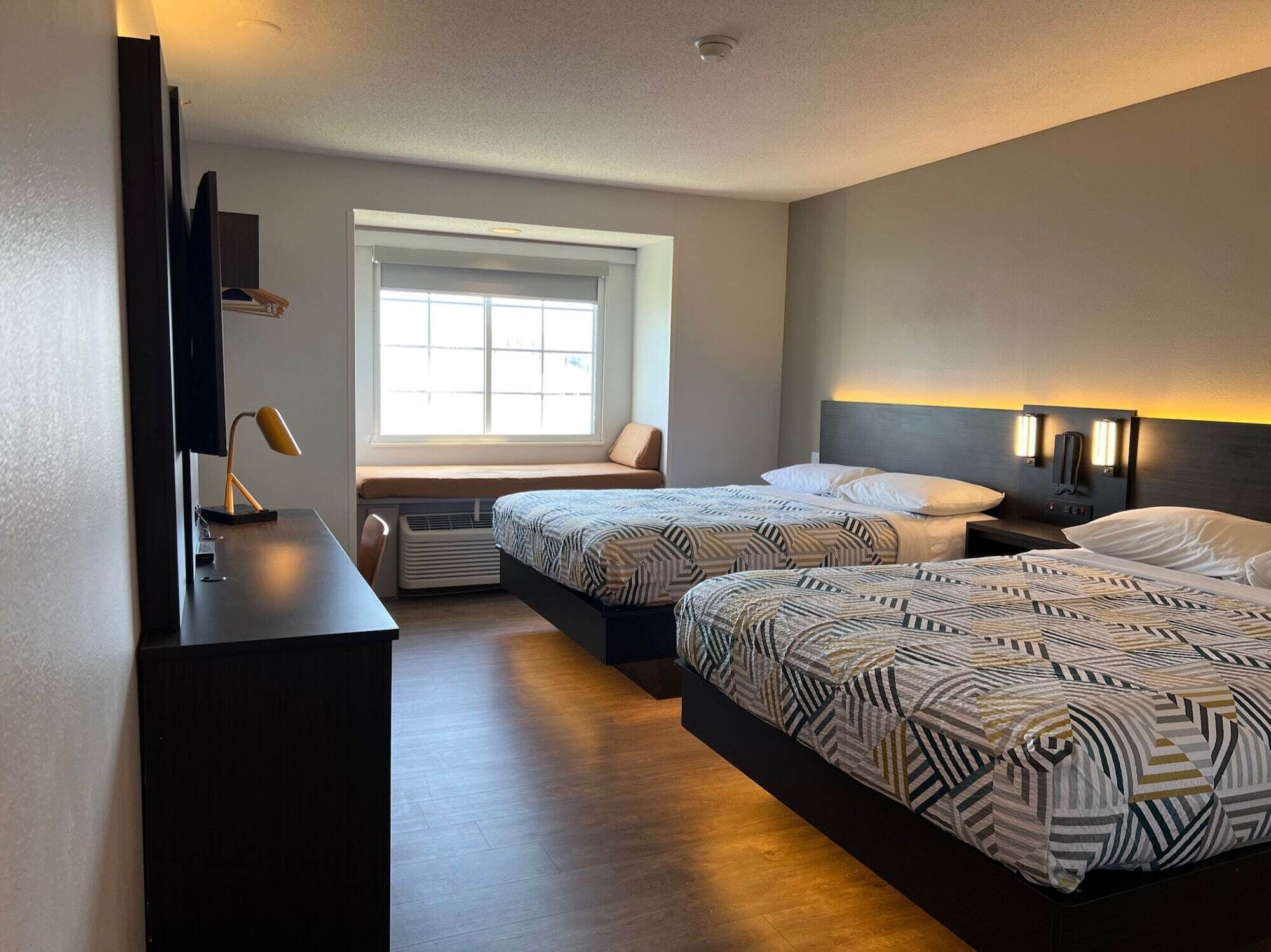 Une chambre d’hôtel avec deux lits, un bureau, une chaise, une fenêtre et un téléviseur fixé au mur.