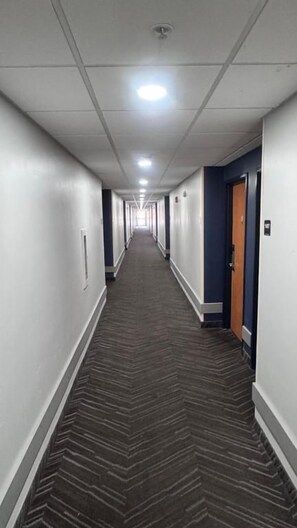 Corredor comprido e estreito de um hotel, com carpete estampado, paredes brancas e portas em ambos os lados.
