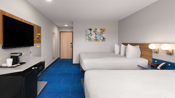 A hotel room with a flat-screen TV, a desk with a coffee maker, two beds, a painting on the wall, and a blue carpet. - Microtel Inn & Suites by Wyndham Independence (Independence)