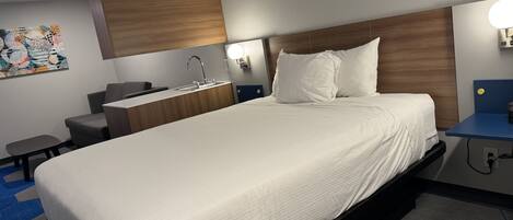 A hotel room with a large bed, a bedside table, a desk, a chair, a small table, a sink, and a wall-mounted light.