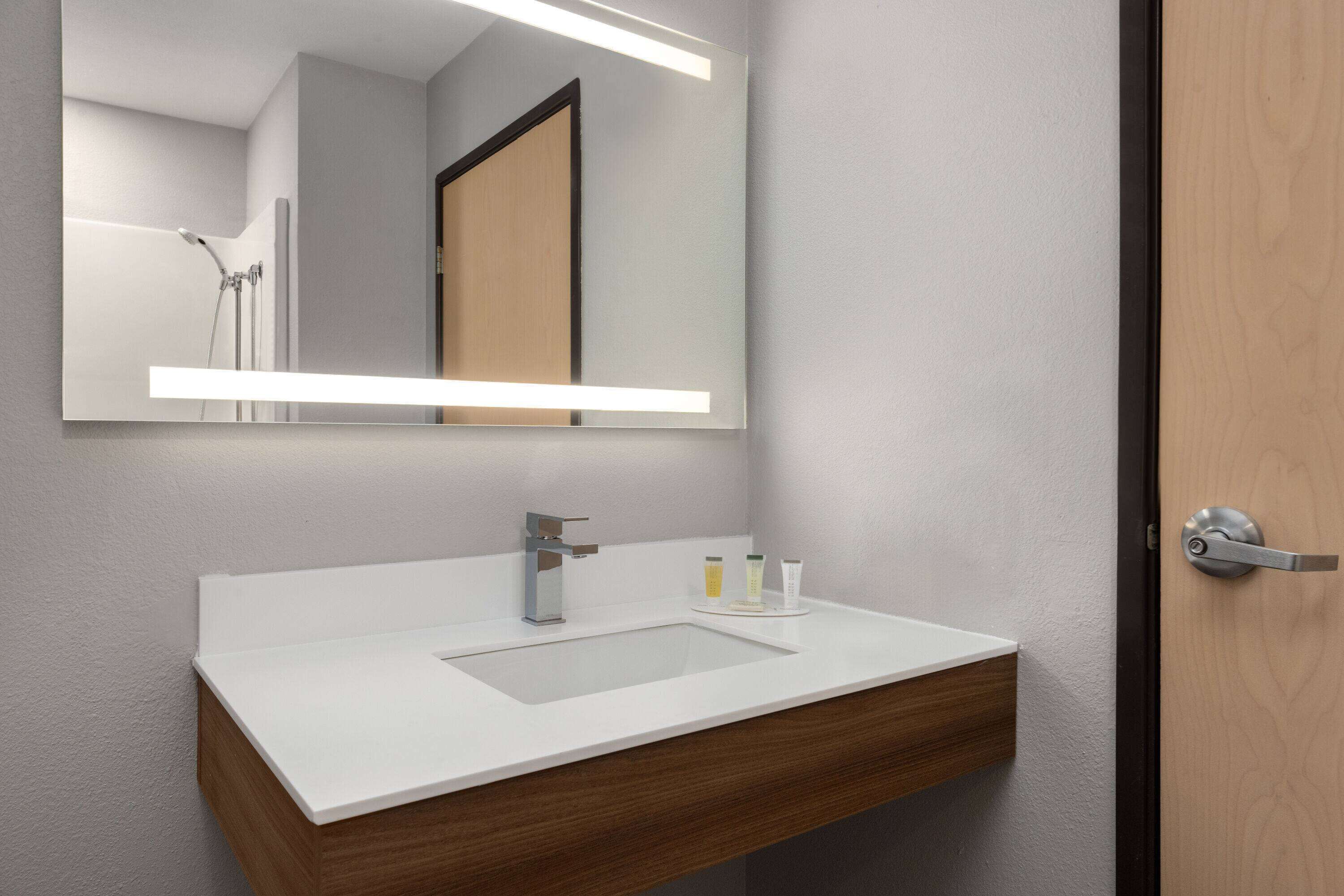 A modern bathroom with a white sink, wooden vanity, and a large mirror with integrated lighting.