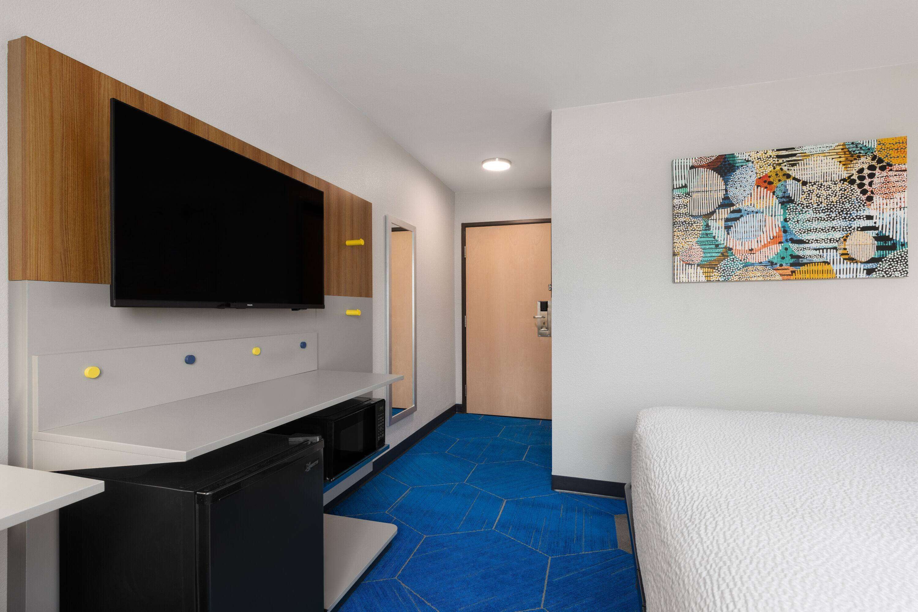A hotel room with a bed, a desk with a flat-screen TV, a microwave, and a door leading to another room.
