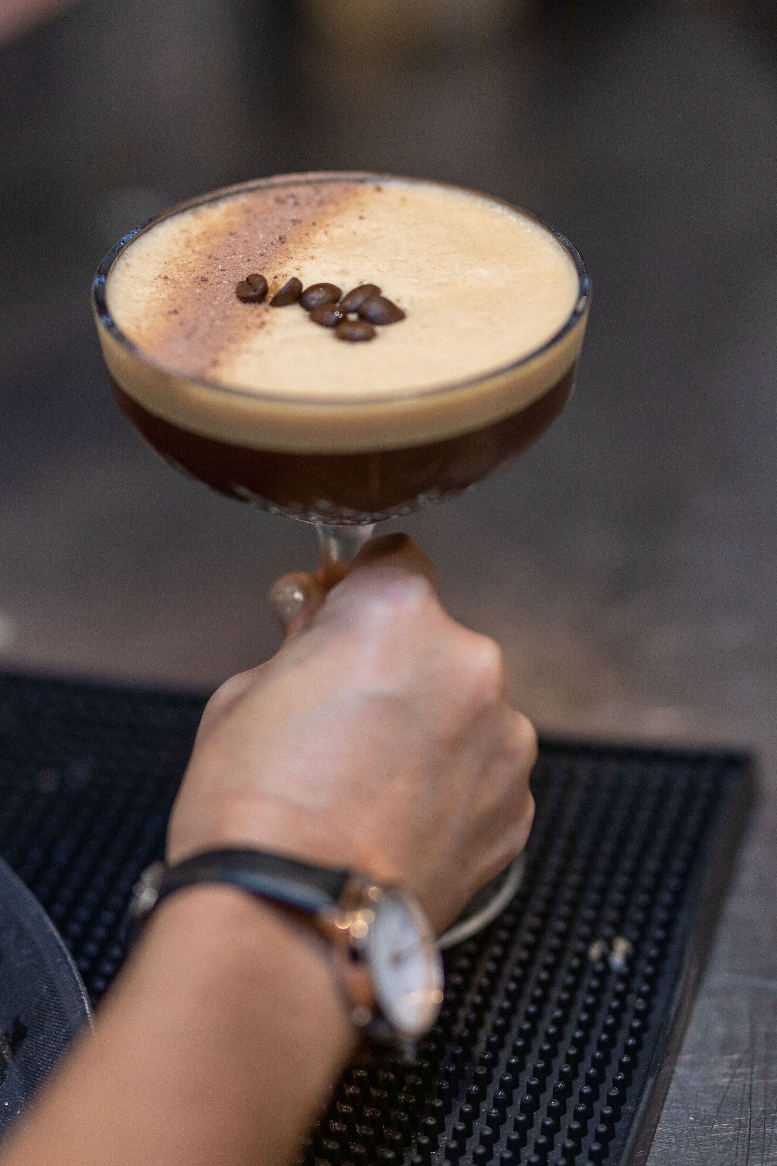A hand holding a cocktail glass with a frothy top and coffee beans on top.