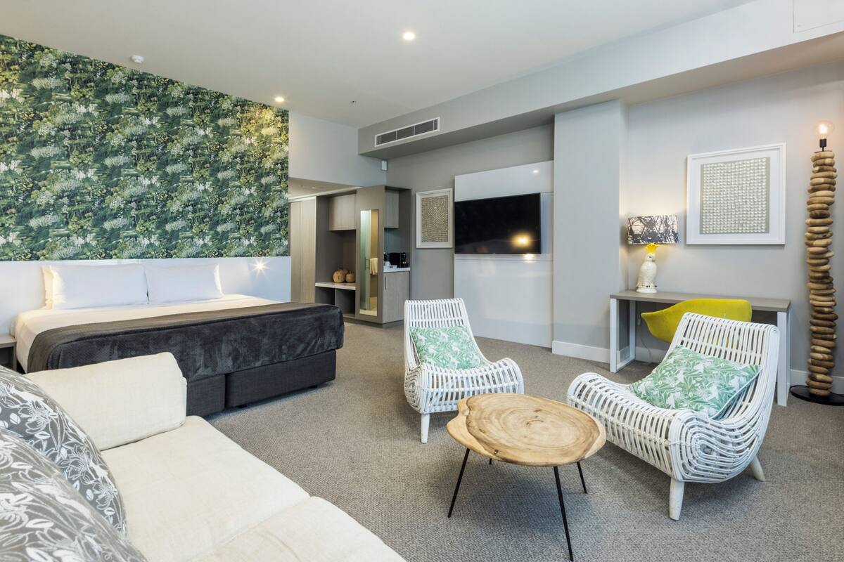 A modern hotel room with a bed, two white wicker chairs, a small wooden table, a desk with a lamp, and a large green patterned wall.