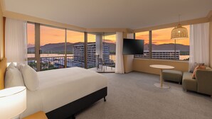 Habitación de hotel con una cama grande, un sofá, un televisor y un balcón con vista a la ciudad y las montañas.