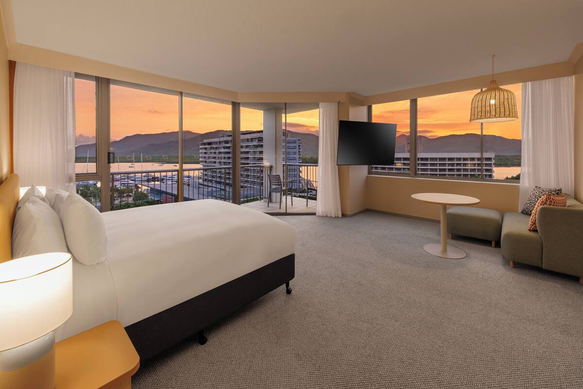 A hotel room with a large bed, a sofa, a TV, and a balcony with a view of the city and mountains.