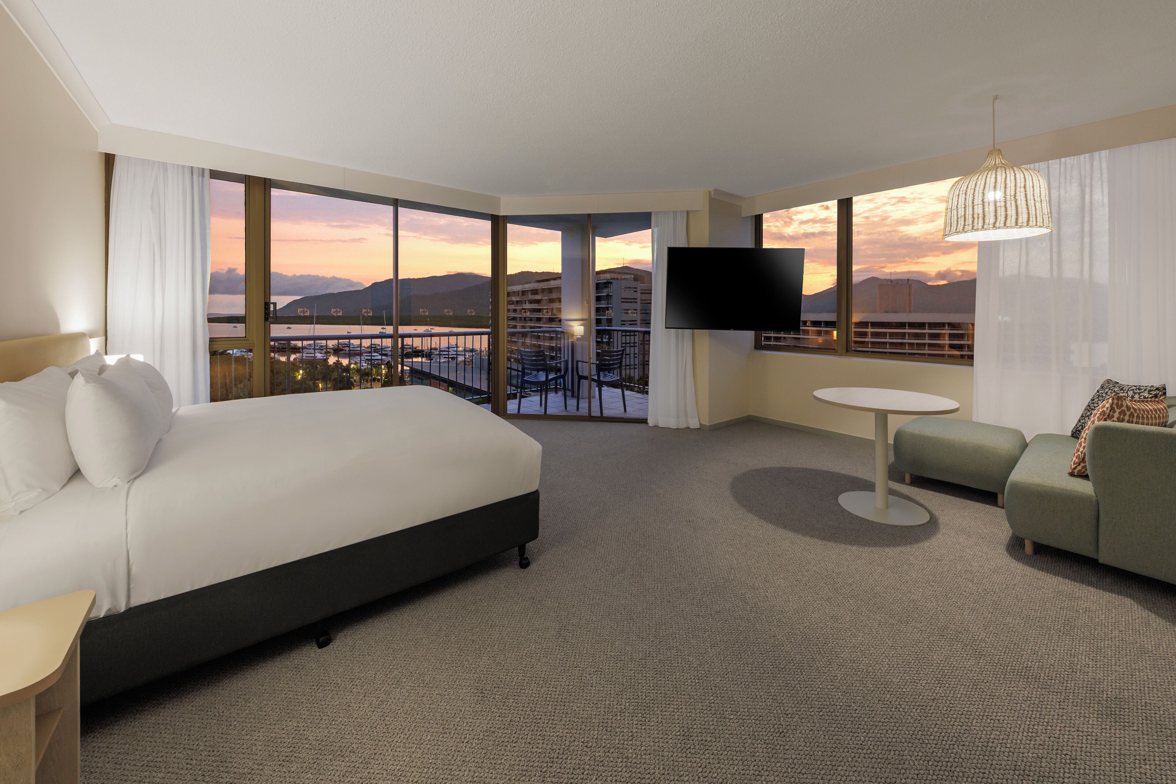 A hotel room with a large bed, a small sofa, a round table, and a TV. The room has large windows with a view of a cityscape and mountains.