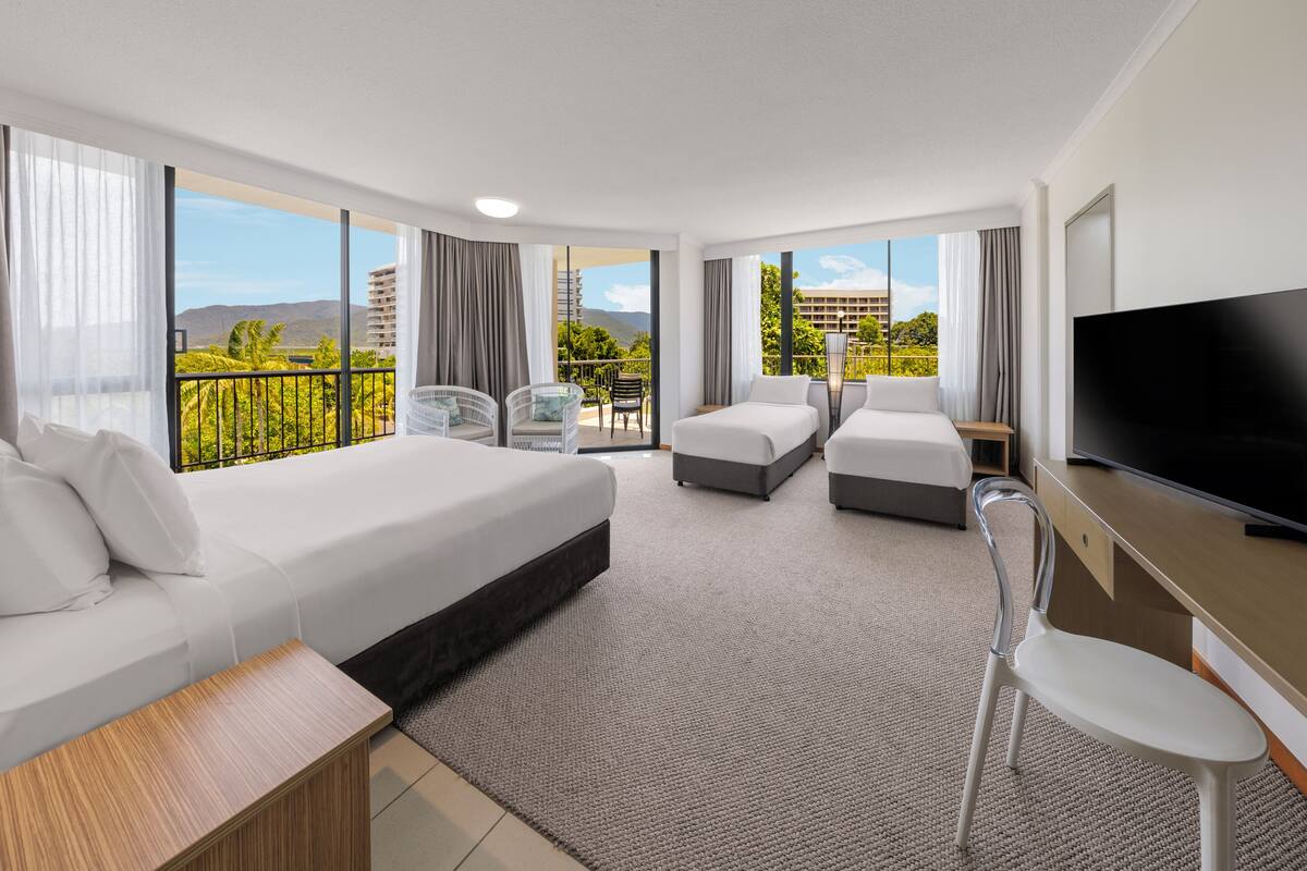 A hotel room with a bed, two smaller beds, a desk, a TV, and a balcony with a view.