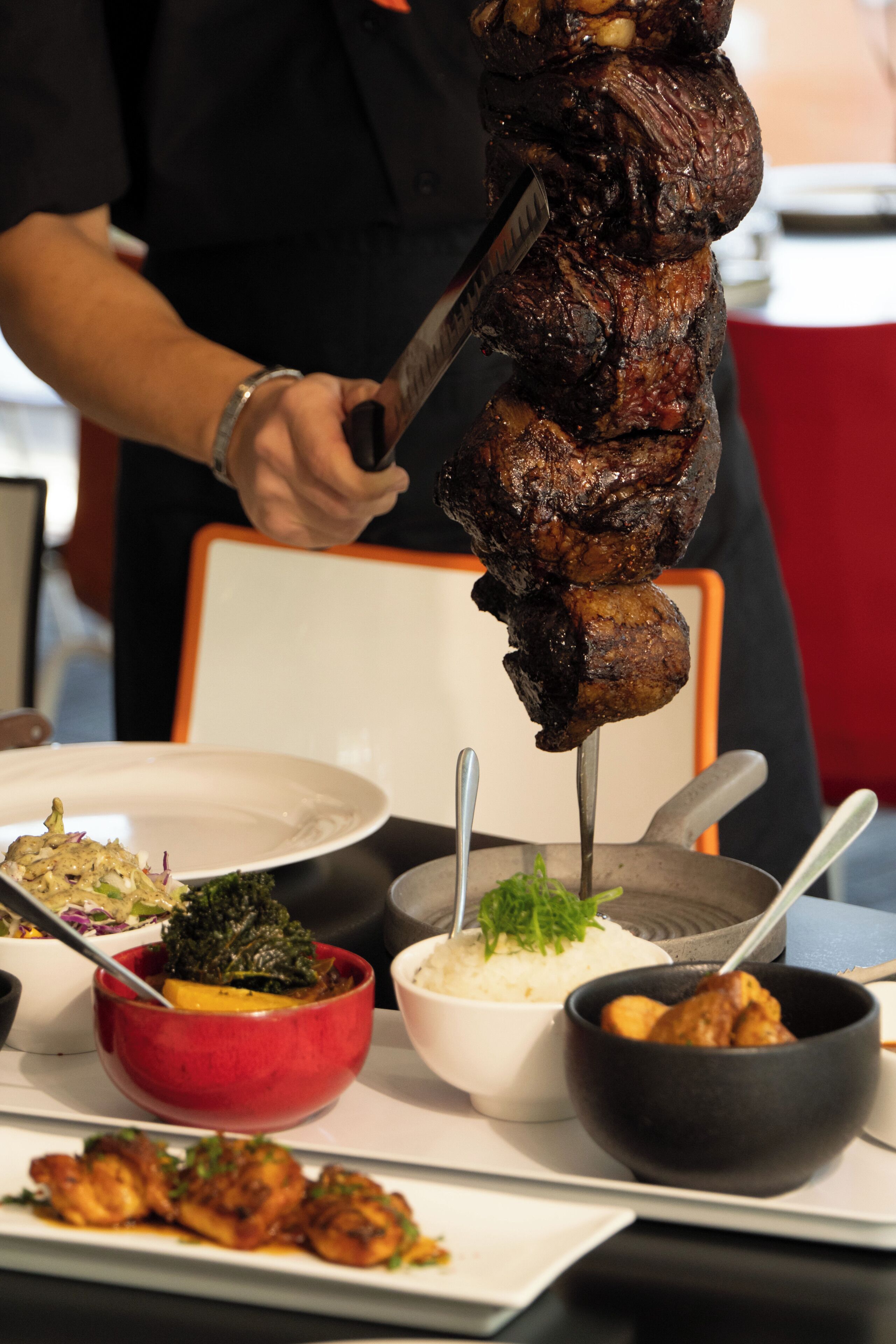 A person carving meat on a vertical rotisserie, surrounded by various dishes including rice, greens, and a red bowl.