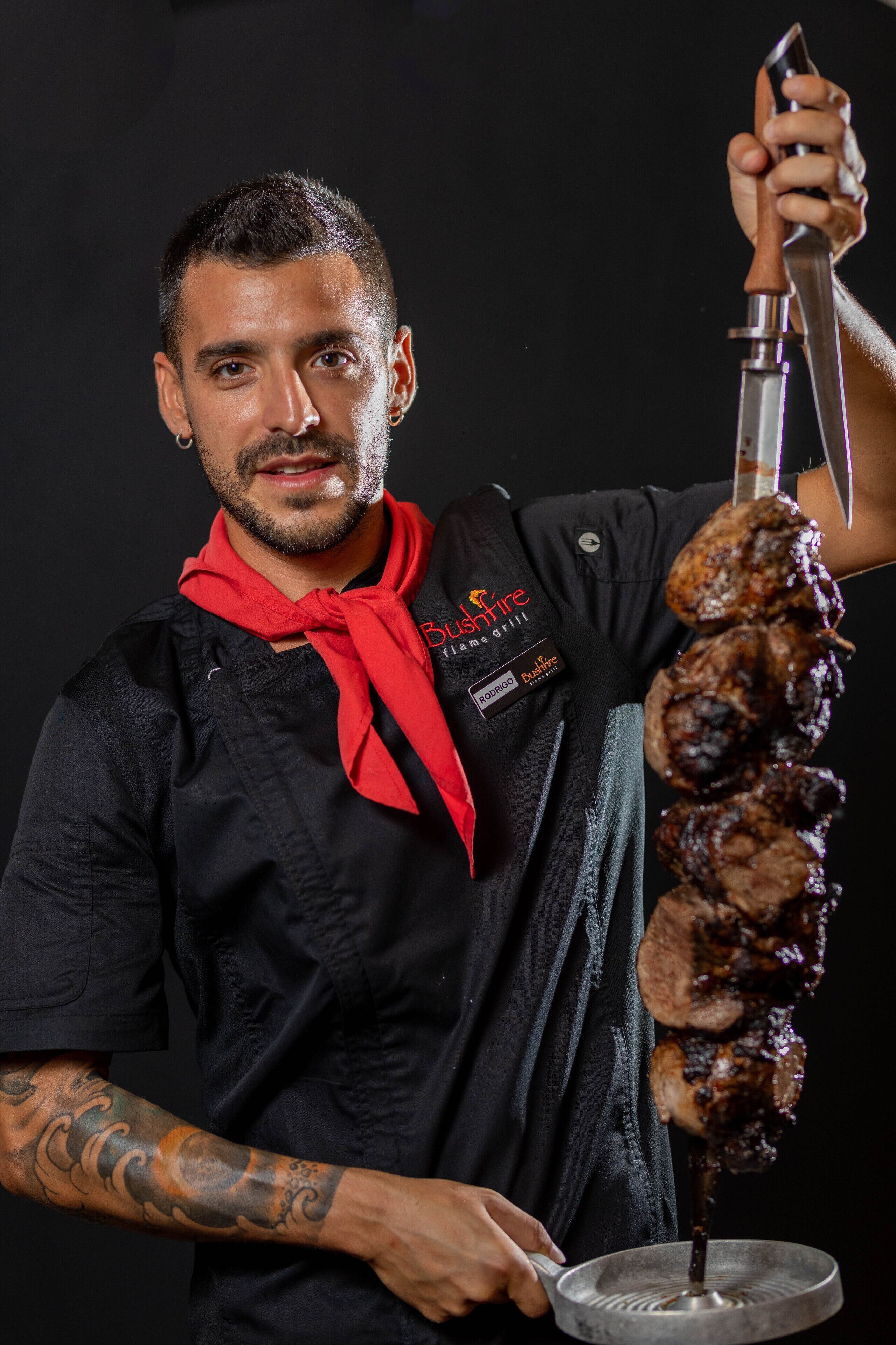 A person holding a large meat skewer with a metal rod, wearing a black shirt with a red scarf and a name tag.