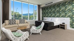 Una habitación de hotel moderna con un gran ventanal, una cama, un sofá, dos sillones de mimbre y una pequeña mesa.
