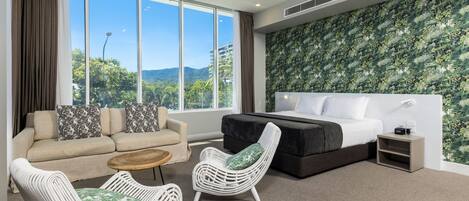 Una habitación de hotel moderna con un gran ventanal, una cama, un sofá, dos sillones de mimbre y una pequeña mesa.