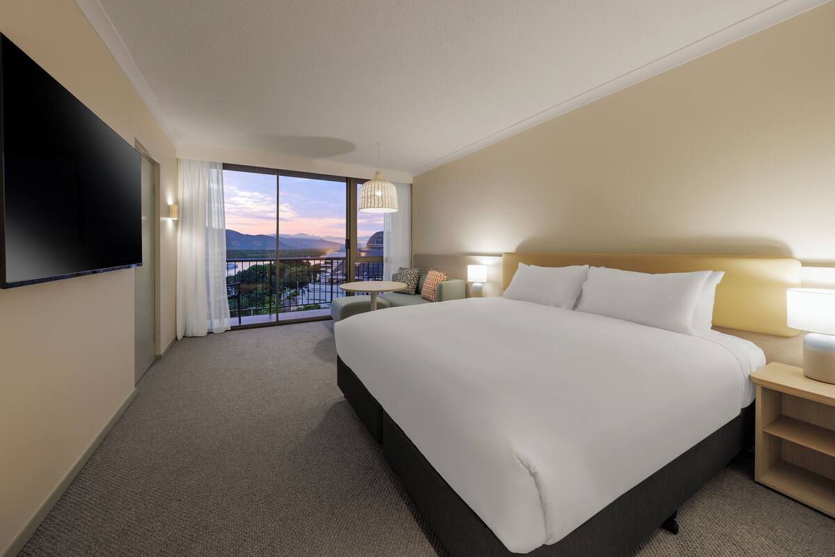A hotel room with a large bed, a flat-screen TV, a balcony with a view of the mountains, and a small table with a lamp.